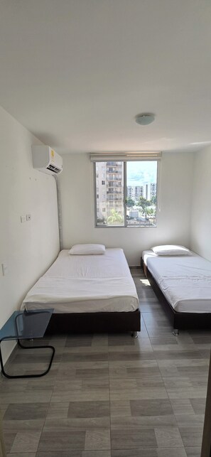 A room with two beds, a window with a view of buildings, an air conditioning unit, and a small table. - Beautiful vacation apartment (Girardot)