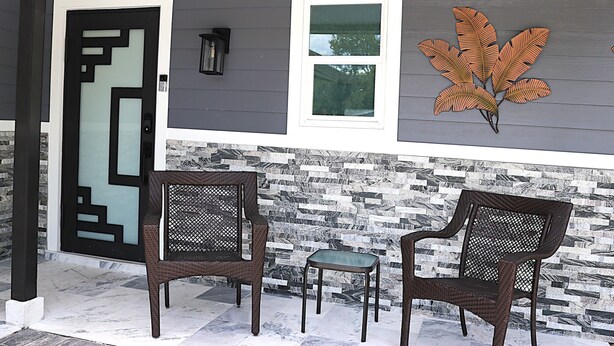 A porch with two wicker chairs and a small table, a decorative leaf wall art, a door with a geometric design, and a window with a view of trees. - Sarasota Silver Oasis - Private Pool, Near Beaches & Downtown (Sarasota)
