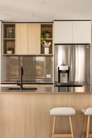 Une cuisine moderne avec des meubles en bois, un réfrigérateur en acier inoxydable, une machine à café et deux tabourets de bar.