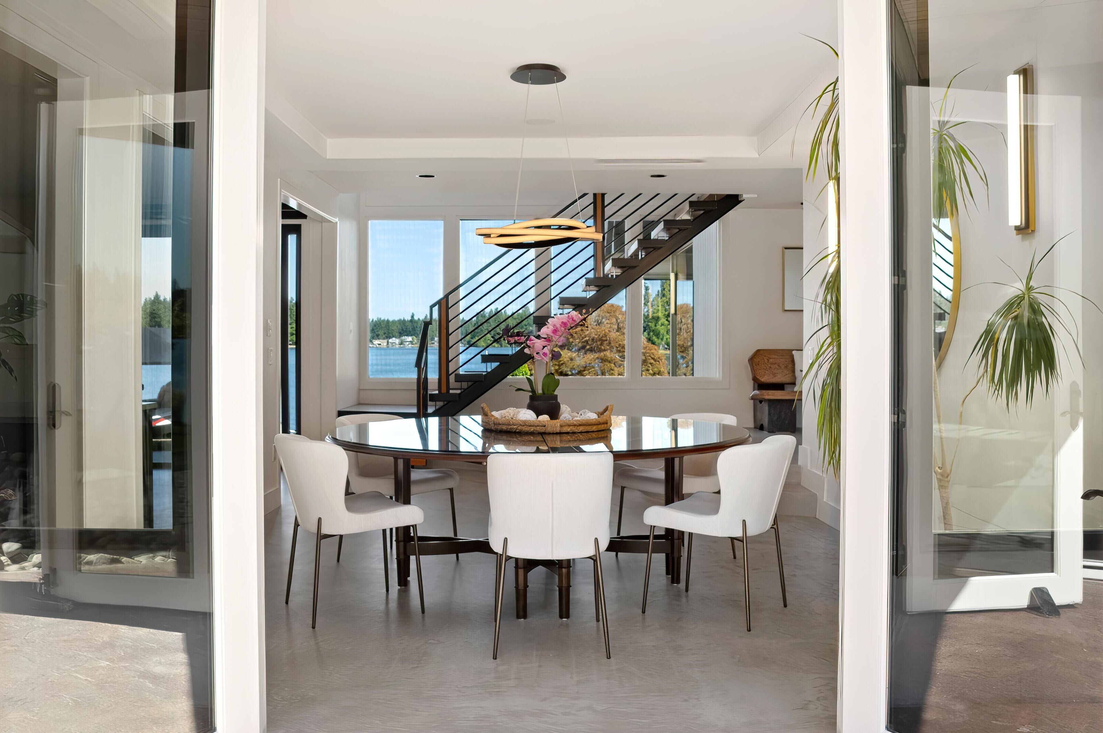 Een moderne eetruimte met een glazen tafel, witte stoelen en een trap naar een balkon.