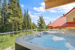 A hot tub with a wooden deck, surrounded by trees and a building with a red roof. - True Ski-In/Out Luxury Chalet • Hot Tub • Sauna (Big White)