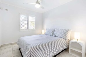 Ein Schlafzimmer mit einem Bett, einem Deckenventilator, einem Fenster mit Jalousien und Nachttischlampen.
