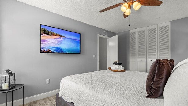 Un dormitorio con una cama, ventilador de techo, televisor en la pared y mesita de noche con una vela.