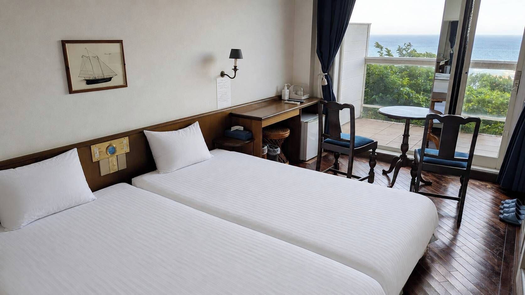 A hotel room with two beds, a desk, a chair, a small table, and a view of the ocean.