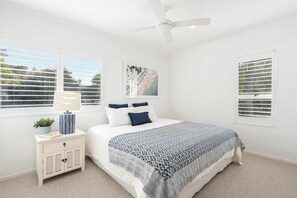 Ein Schlafzimmer mit einem Bett, einem Nachttisch, einem Deckenventilator und zwei Fenstern mit Jalousien.