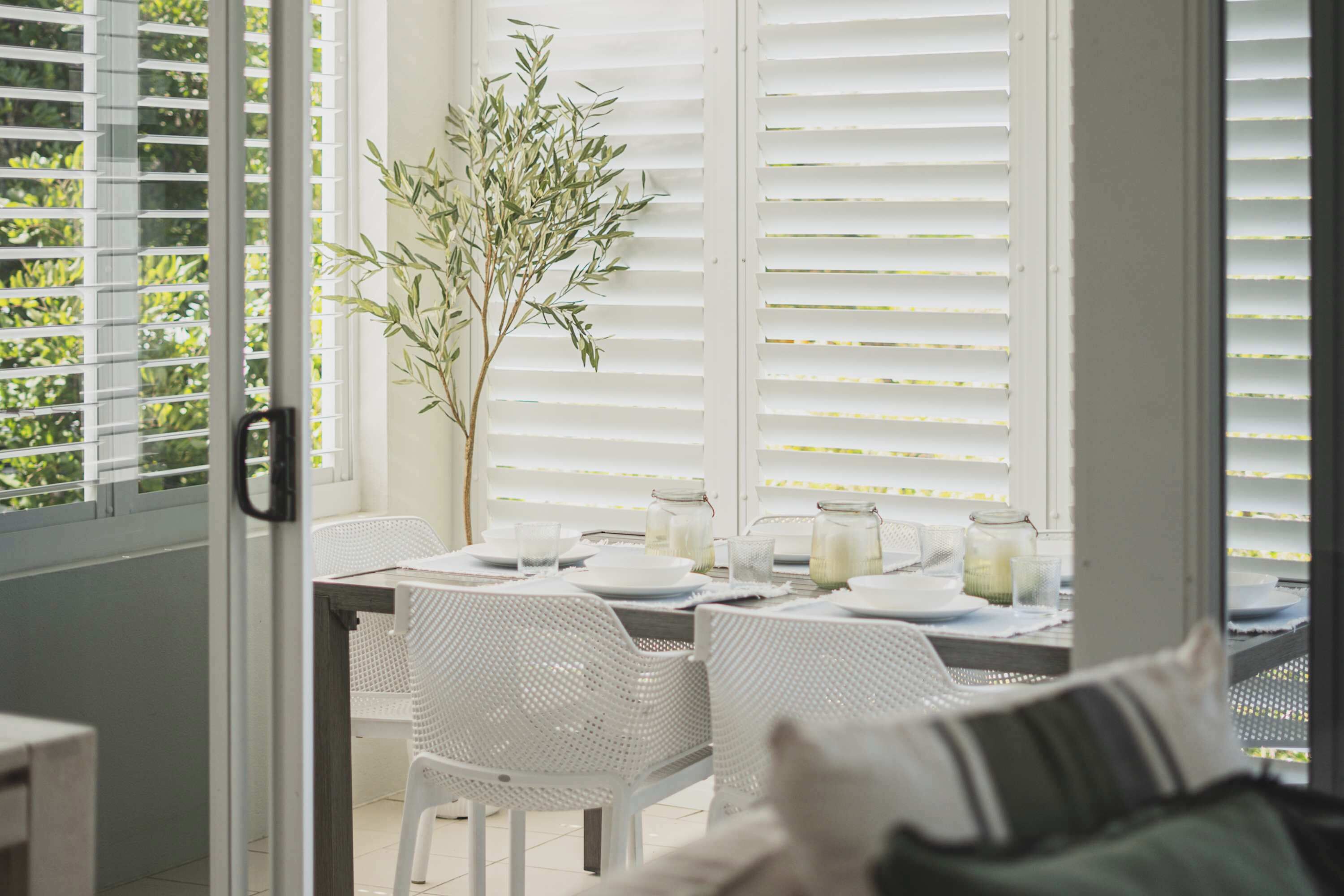 Un coin repas avec une table dressée pour deux personnes, des chaises en osier blanches, une plante en pot et une vue à travers des stores blancs.