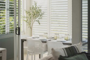 Un coin repas avec une table dressée pour deux personnes, des chaises en osier blanches, une plante en pot et une vue à travers des stores blancs.