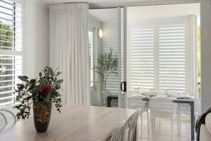 Ein Essbereich mit Holztisch, weißen Stühlen und Blick durch eine Glastür auf einen Außenbereich mit weißen Jalousien.