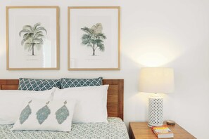 Une chambre à coucher bien rangée, avec un lit, des oreillers, une table de chevet avec une lampe, et deux illustrations botaniques encadrées accrochées au mur.