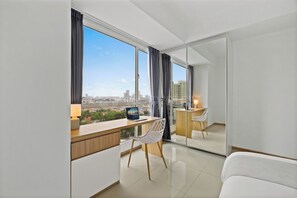 Quarto moderno com vista para a cidade, uma escrivaninha com um laptop, uma cadeira e uma cama.