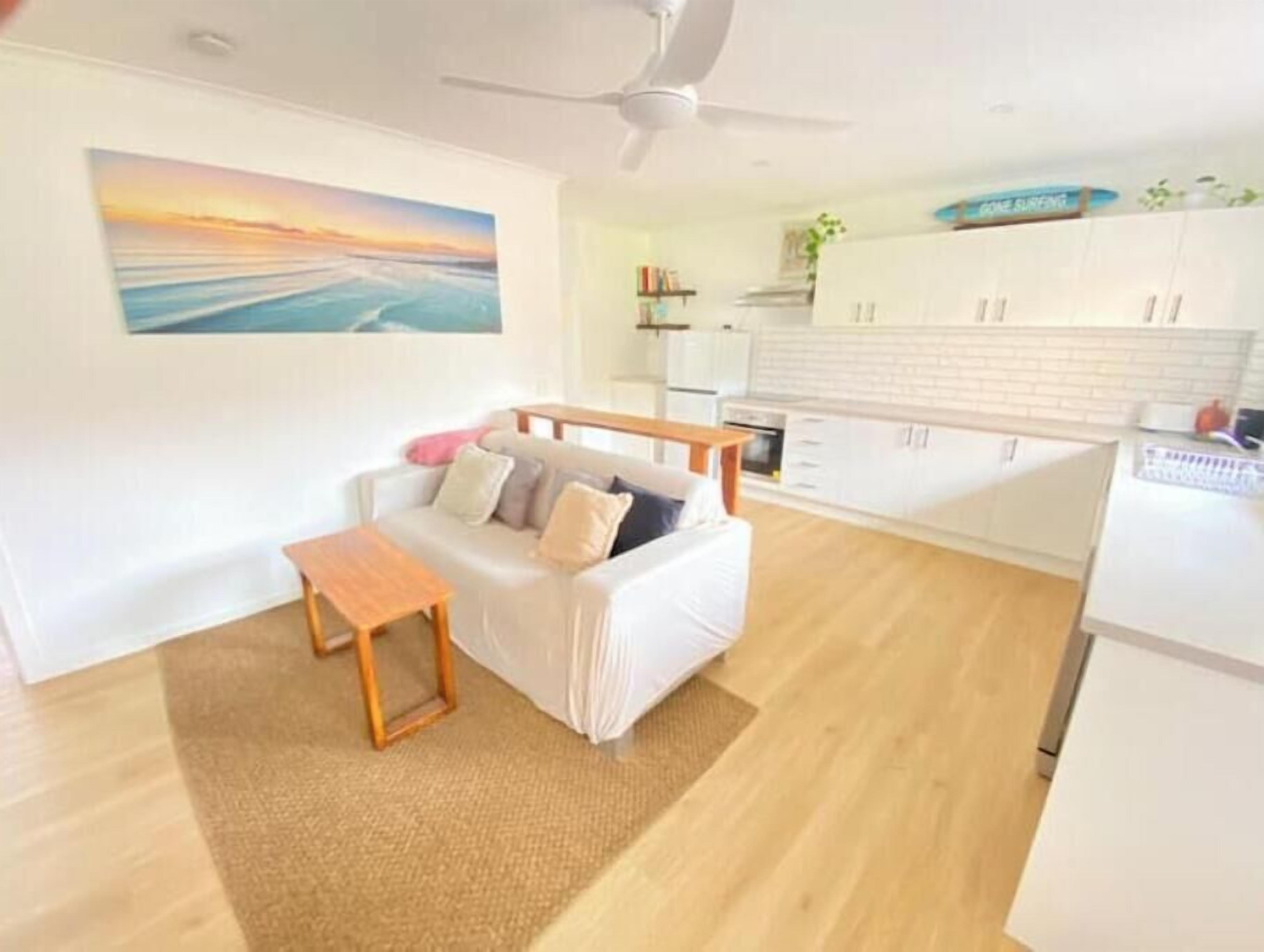 A modern living room with a sofa, a small wooden table, and a ceiling fan. A kitchen area with white cabinets and a refrigerator is visible in the background.