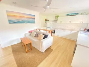 A modern living room with a sofa, a small wooden table, and a ceiling fan. A kitchen area with white cabinets and a refrigerator is visible in the background. - Surf, Beach, Fishing, Fishing, Relaxing, SUP, paddling, Restaurants, cafes (Currumbin)
