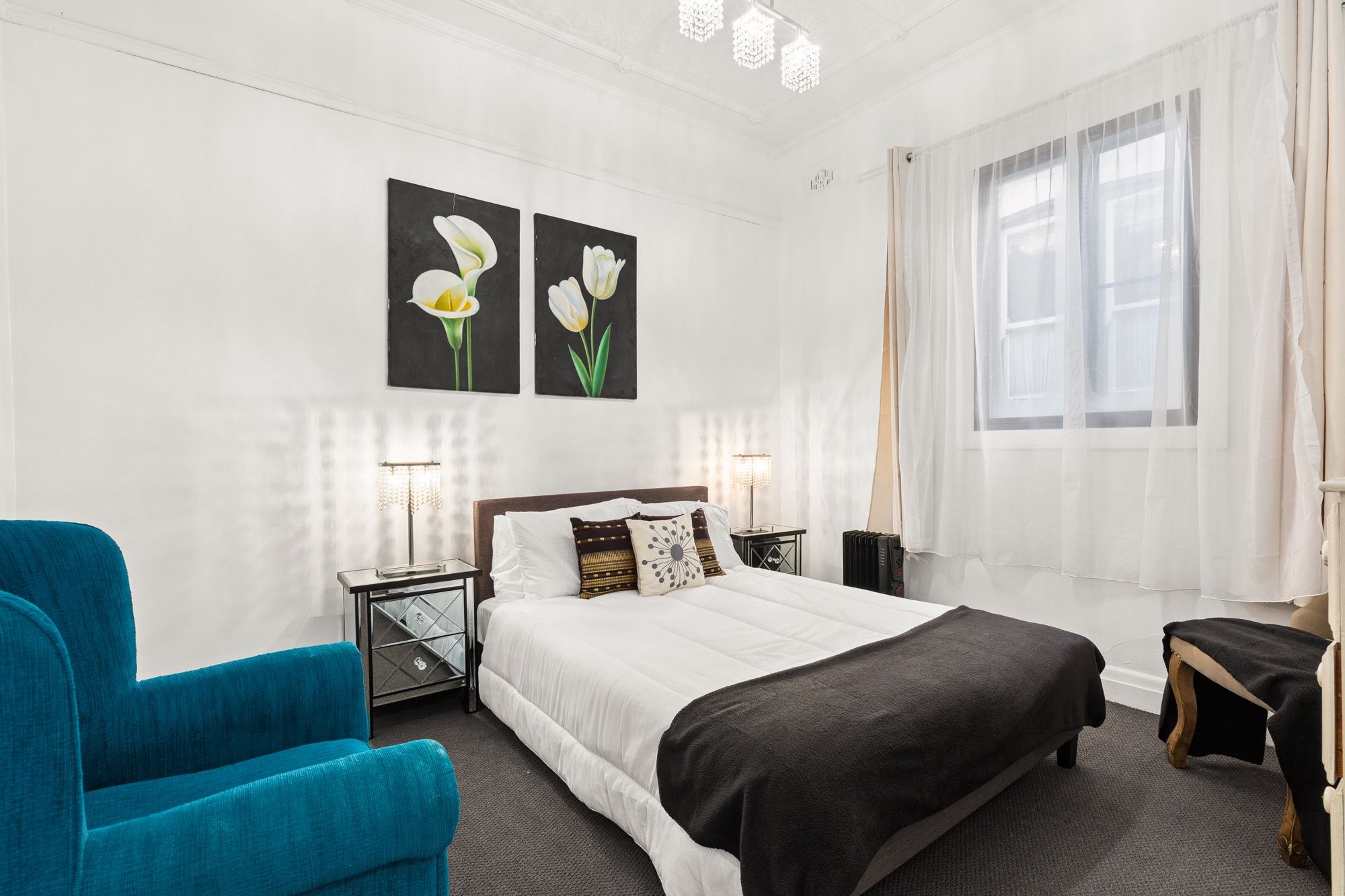 Une chambre d’hôtel avec un lit, un fauteuil bleu, une petite table et deux lampes fixées au mur.