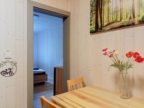Een houten interieur met een eettafel, stoelen, een vaas met bloemen, een aan de muur hangend schilderij en een deur naar een andere kamer.