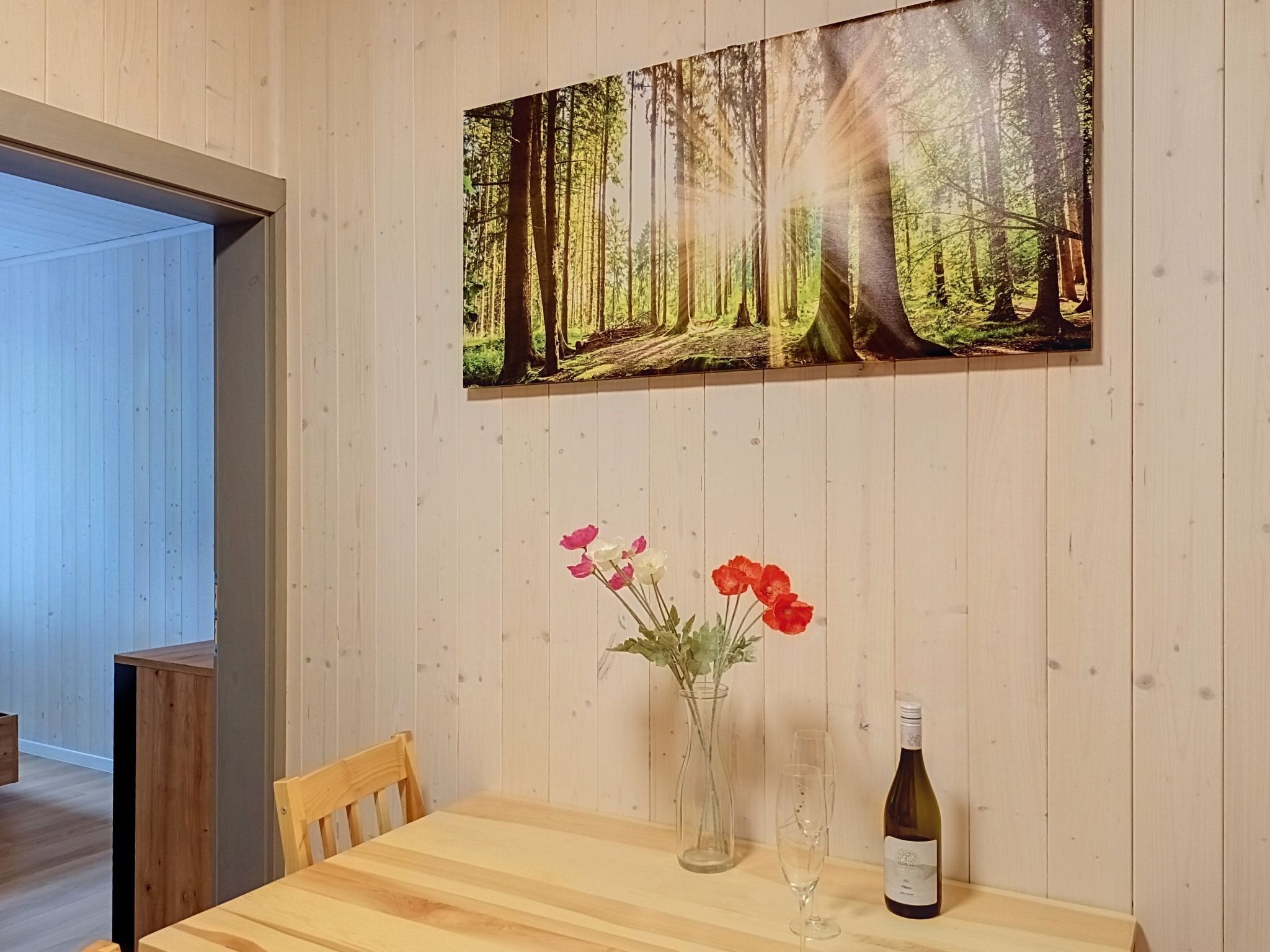 Ein Holztisch mit einem Waldbild, einer Blumenvase und einer Weinflasche.