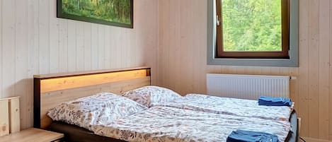 A wooden-paneled room with a bed, a nightstand, a window with a view of trees, and a painting on the wall.
