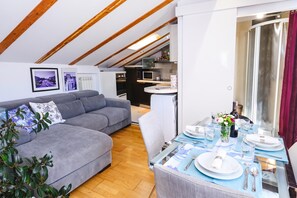 Ein modernes Wohnzimmer mit einem grauen Ecksofa, einem Esstisch für zwei Personen und einer Küche im Hintergrund.