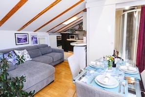 A modern living room with a grey sectional sofa, a dining table set for two, and a kitchen in the background. - Casa Terrazza in Historic Koper With Sea View (Koper)