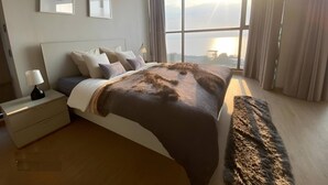 A modern bedroom with a large bed, a nightstand, a lamp, and a view of the city through large windows. - Sea View Large Luxury Space Beach Front (Pattaya)