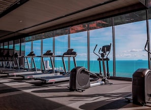 A gym with treadmills, ellipticals, and a stationary bike, with a view of the ocean and buildings through large windows. - Sea View Large Luxury Space Beach Front (Pattaya)