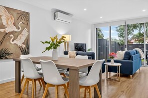 A modern living room with a dining area, featuring a large painting, a dining table with chairs, a sofa, and a small side table with a lamp. - The Garfield Nest — Steps From Everything (Kurralta Park)