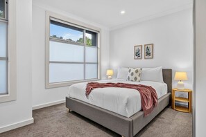 A bedroom with a large window, a bed with a grey headboard, two bedside lamps, and two framed pictures on the wall. - The Garfield Nest — Steps From Everything (Kurralta Park)