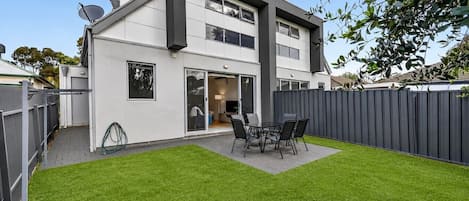 A modern two-story house with a flat roof, a covered patio with a table and chairs, and a well-maintained lawn.