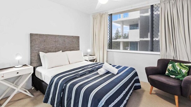 A hotel room with a bed, a desk, a chair, a ceiling fan, and a window with curtains. - Iconic Burleigh Beachfront, 2 Bedroom Apartment (Gold Coast)
