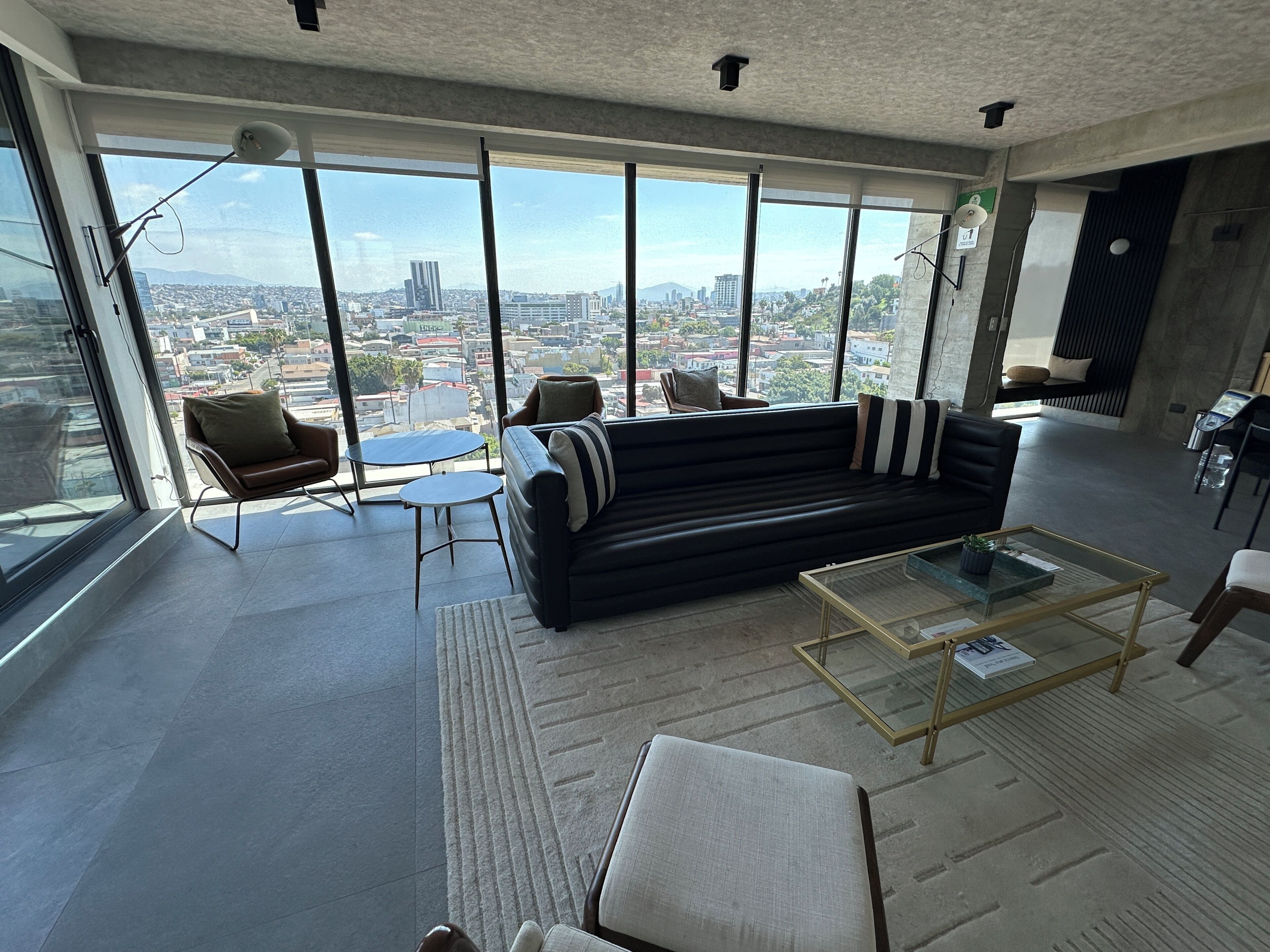 A modern living room with a black sofa, a glass coffee table, and a view of a cityscape through large windows.