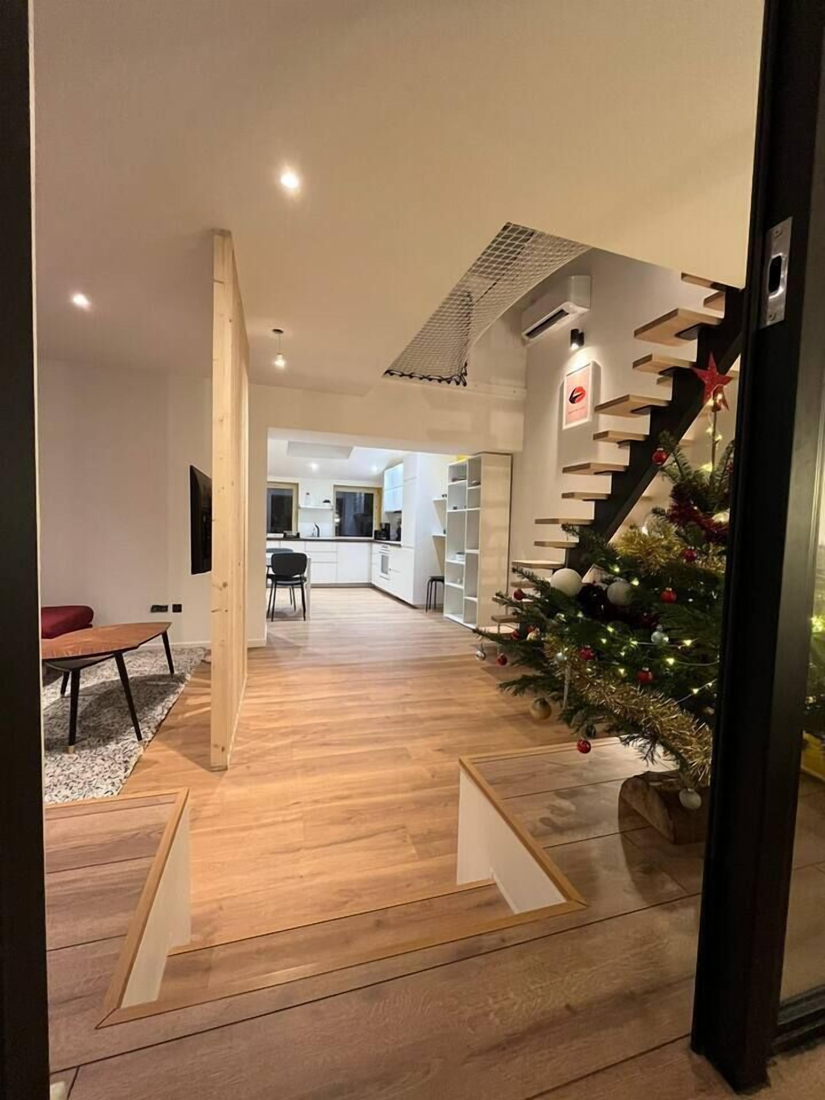 A modern interior with a wooden floor, a staircase with a Christmas tree, and a kitchen area in the background.
