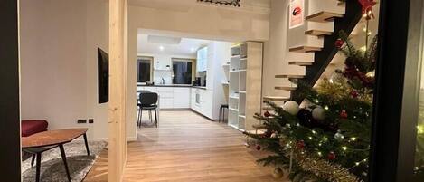 Un interior moderno con suelo de madera, una escalera con un árbol de Navidad y una zona de cocina al fondo.