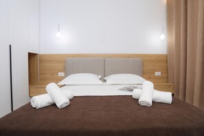 A neatly made bed with two pillows and two rolled towels, a wooden headboard, and two wall-mounted lights. - Sea Side Palm Modern Suites (Orikum)