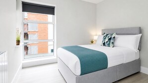 A modern bedroom with a bed, bedside table, lamp, and a view of a building through a window. - Modern Central Dublin Apartment  Near Bolton Square (Dublin 1)