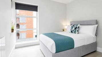 Un dormitorio moderno con cama, mesita de noche, lámpara y vista a un edificio a través de una ventana.