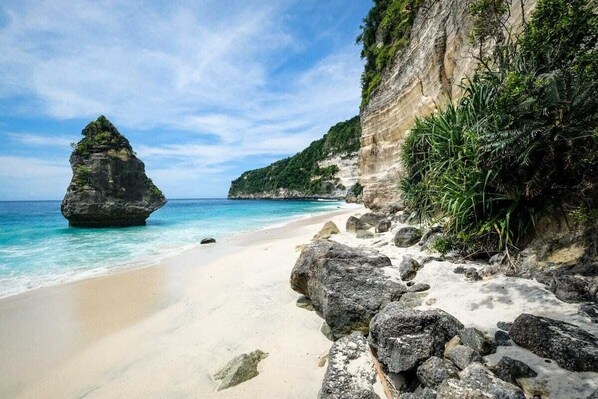 A beach with clear turquoise water, a large rock formation, and a cliff with vegetation.