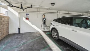 Ein weißes Auto steht in einer überdachten Parkgarage, mit einer Ladestation, einem 'Verbotenes Parken'-Schild und einem Warnhinweis an der Wand.