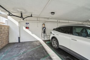 A white car parked in a covered parking area with a charging station, a 'No Parking' sign, and a 'Warning' sign on the wall. - Lovely 1-bedroom condo in Carpinteria steps from the beach (Carpinteria)