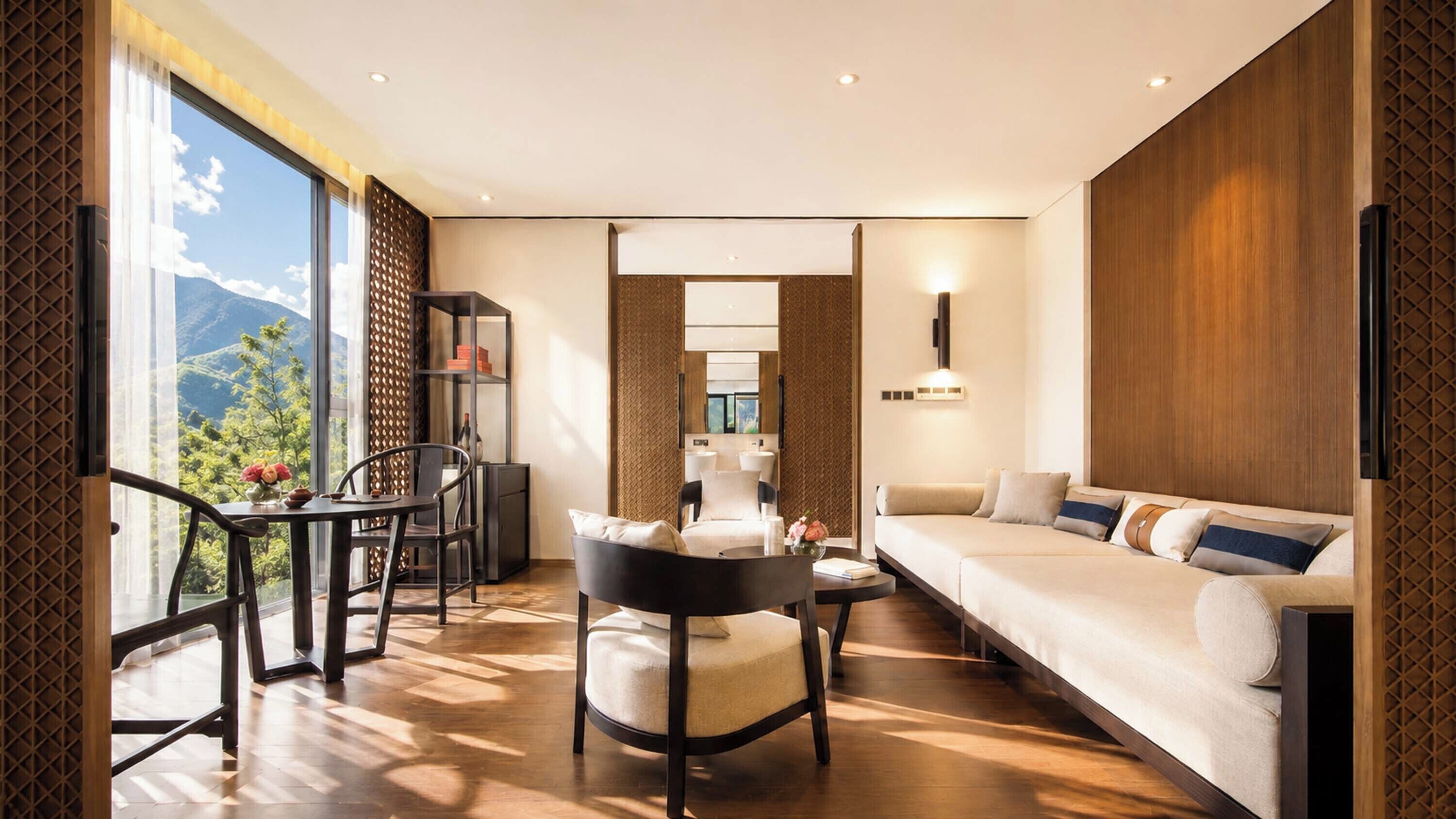 Ein modernes Hotelzimmer mit einer Couch, einem Esstisch mit Stühlen, einem kleinen Tisch mit einer Vase und Bergblick durch große Fenster.