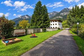 Ein Weg führt zu einem weißen Gebäude mit zahlreichen Fenstern, umgeben von Grünflächen und Bergen im Hintergrund.