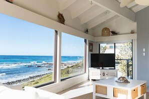 Ein modernes Wohnzimmer mit großem Fenster mit Blick auf eine zerklüftete Küstenlinie, einem Flachbildfernseher und einem weißen Konsolentisch.