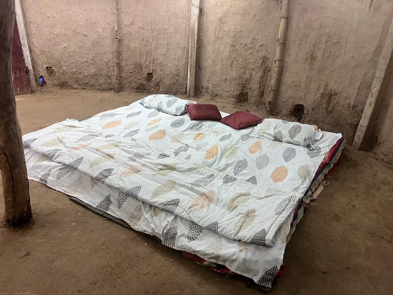 A single bed with a patterned bedspread in a room with concrete walls and a dirt floor.