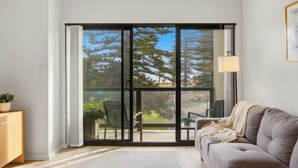 A room with a sliding glass door leading to an outdoor area, a sofa, a small table, and a lamp. - Belle Escapes - Bayview Escape Glenelg (Holdfast Bay)
