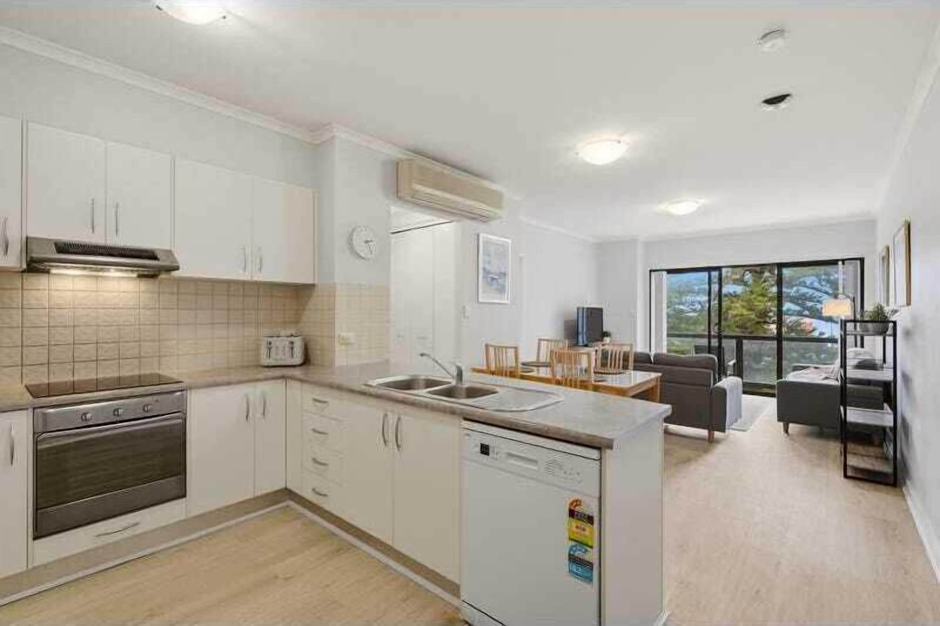 A modern kitchen with white cabinets, a built-in oven, and a dishwasher. There is a dining area with a table and chairs, and a large window providing a view of the outdoors.