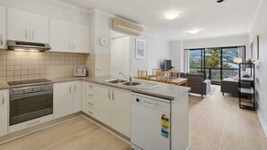A modern kitchen with white cabinets, a built-in oven, and a dishwasher. There is a dining area with a table and chairs, and a large window providing a view of the outdoors. - Belle Escapes - Bayview Escape Glenelg (Holdfast Bay)