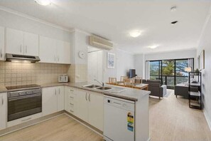 Eine moderne Küche mit weißen Schränken, einem eingebauten Backofen und einem Geschirrspüler. Es gibt eine Essecke mit Tisch und Stühlen sowie ein großes Fenster mit Blick ins Freie.