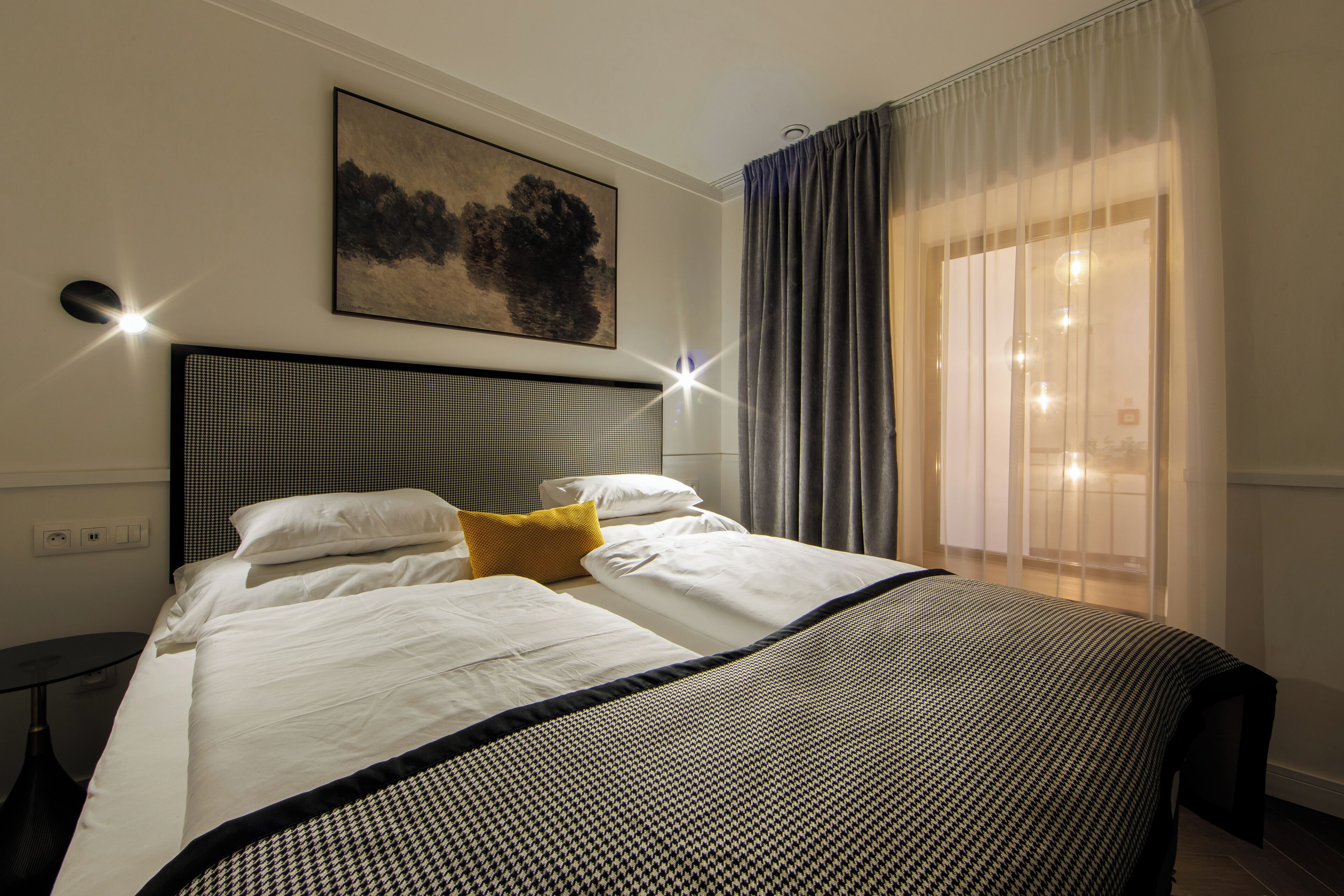 Quarto de hotel com uma cama, cabeceira, pintura na parede, janela com cortinas e mesa de cabeceira.
