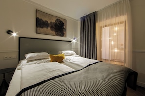 Una habitación de hotel con una cama, cabecera, un cuadro en la pared, una ventana con cortinas y una mesita de noche.
