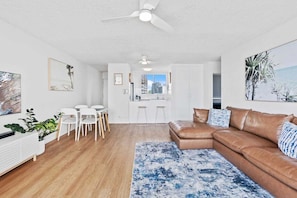A modern living room with a brown leather sofa, a dining area with white chairs, and a kitchen in the background. - Escape on the Esplanade (Gold Coast)