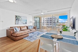 A modern living room with a brown leather sofa, a dining table with chairs, and a large window overlooking a building. - Escape on the Esplanade (Gold Coast)