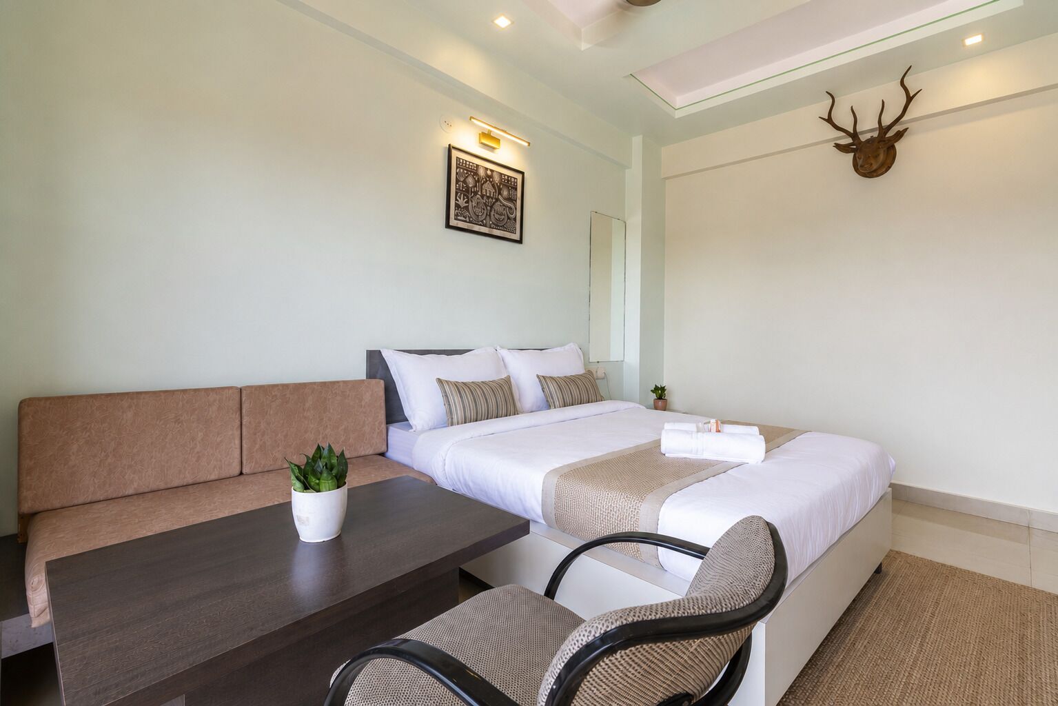 Habitación de hotel con cama, sofá, mesa de comedor y una silla. Hay una planta sobre la mesa y un espejo montado en la pared.